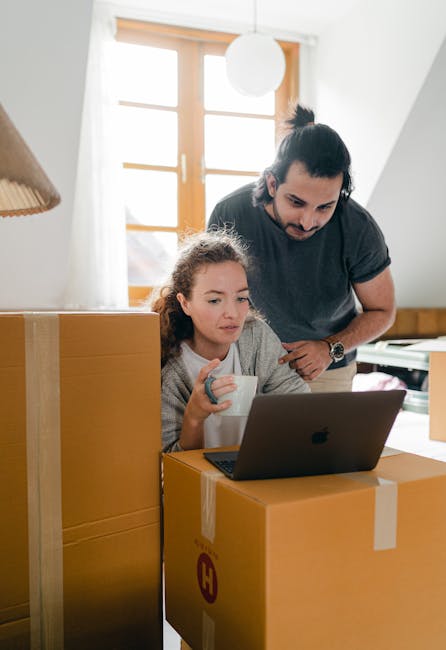 A man and a young girl are inside a home, engaged in house removal activities related to furniture transport and packing. The girl is seated in front of an open laptop resting on a stack of large, sealed cardboard boxes, holding a white mug. The man stands behind her, leaning slightly forward, observing the laptop screen, with his hands resting on a box. The scene takes place near a window with wooden frames and white curtains, allowing natural light to illuminate the space. The room contains other packing materials, such as additional cardboard boxes, and a wooden sideboard is visible in the background. The environment suggests preparation for a house relocation, with the furniture and boxes indicating an ongoing moving process supported by [COMPANY_NAME], Man With a Van Hounslow, specializing in removals and relocation services.