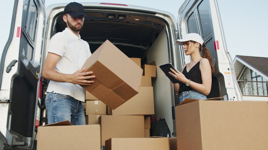 A man and a woman are engaged in a home relocation process involving a large white moving van parked outside a residential property. The man, dressed in a white t-shirt, jeans, and a dark cap, is inside the van, holding a cardboard box filled with various packed items, preparing for furniture transport or loading. The woman, wearing a white cap, a black sleeveless top, and blue jeans, is standing nearby on the pavement, using a tablet or mobile device, possibly coordinating the packing and moving process. The interior of the van contains multiple cardboard boxes of different sizes, some stacked on top of each other, along with packing materials such as bubble wrap and plastic covers for furniture. The environment is well-lit with natural daylight, and part of the house with a balcony and window is visible in the background, indicating the scene takes place during a home move or removal. Man With a Van Hounslow performs services related to removals, including packing, transporting, and logistics for house relocations.
