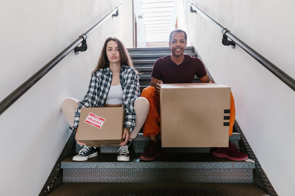 A photograph of two movers from Man With a Van Hounslow positioned on a staircase during a home relocation process. The woman on the left, with long brown hair, is wearing a plaid shirt over a white top and is seated on the stairs holding a small cardboard box labeled 'Fragile.' She is wearing dark sneakers with white laces. The man on the right, with short curly hair, is wearing a maroon t-shirt and orange pants, kneeling next to a larger cardboard box, also held on his lap. Both are on a metal staircase with black railings, inside a well-lit building with light-colored walls and a window at the top of the stairs providing natural light. The scene depicts the careful handling and loading of packing materials and furniture, illustrating the logistics involved in furniture transport and packing for a professional household removal, supported by the expertise of Man With a Van Hounslow, known for their reliable removal services.