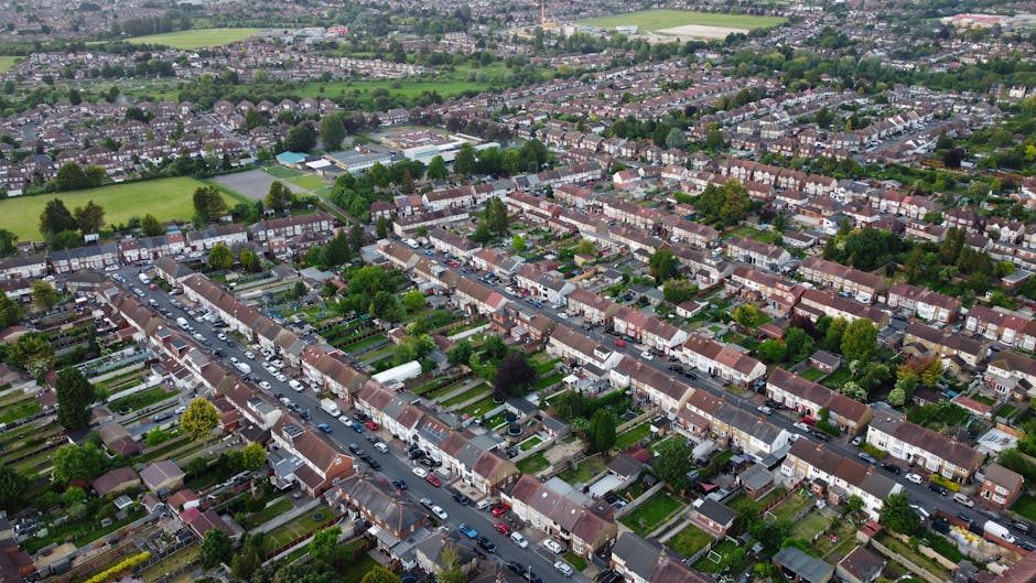 An aerial view of a suburban residential area in Hounslow showing a row of terraced houses with front gardens and driveways, parked cars along the street, and a mix of trees lining the pavement. In the background, green fields and open spaces are visible, with some larger buildings indicating community or recreational facilities. The scene captures the typical layout of a UK home environment during daylight hours, with natural lighting highlighting the rooftops, green foliage, and the movement of vehicles as part of the home relocation or furniture transport process, illustrating common access points and the environment involved in house removals facilitated by Man With a Van Hounslow.