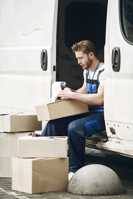 A photograph of two movers from Man With a Van Hounslow positioned on a staircase during a home relocation process. The woman on the left, with long brown hair, is wearing a plaid shirt over a white top and is seated on the stairs holding a small cardboard box labeled 'Fragile.' She is wearing dark sneakers with white laces. The man on the right, with short curly hair, is wearing a maroon t-shirt and orange pants, kneeling next to a larger cardboard box, also held on his lap. Both are on a metal staircase with black railings, inside a well-lit building with light-colored walls and a window at the top of the stairs providing natural light. The scene depicts the careful handling and loading of packing materials and furniture, illustrating the logistics involved in furniture transport and packing for a professional household removal, supported by the expertise of Man With a Van Hounslow, known for their reliable removal services.