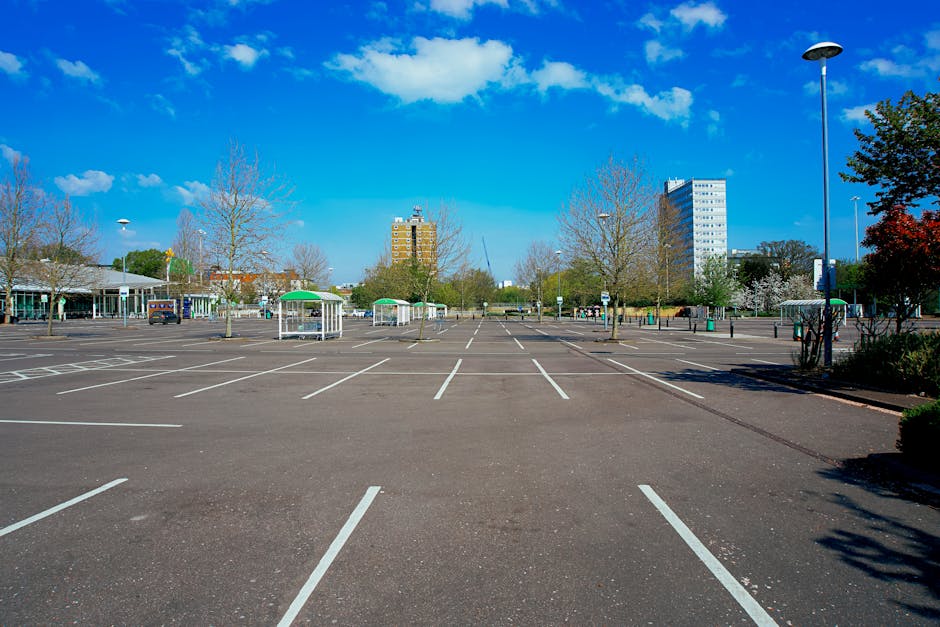 An empty parking lot situated in an urban area during daytime, with clear blue skies and scattered white clouds overhead. The asphalt surface of the parking area is marked with white lines designating individual parking spaces, some of which are occupied by small metal and glass bus shelters with green roofs positioned near the center and sides. Leafless trees line the perimeter of the lot, with a few bearing early spring blossoms, and tall lampposts with black fixtures are visible along the edges of the parking area. In the background, modern office or apartment buildings with large windows and balconies rise above the trees, indicating a city center setting. The parking lot is well-lit by natural sunlight, illustrating a typical scene where home relocation or furniture transport might involve careful planning for parking permissions, as managed by local authorities such as Hounslow Council, with services provided by companies like Man With a Van Hounslow for moves requiring parking permits and logistical support.