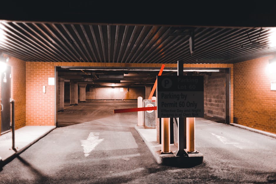 A parking garage entrance featuring a black overhead ceiling with linear wooden panels, flanked by brick walls on either side. In the foreground, a sign indicating parking regulations for permit holders is mounted on a black pole, with orange and red barriers halting vehicle access. The barriers are attached to short concrete posts, positioned on a paved surface with a white directional arrow painted on the ground pointing towards the garage interior. Inside, the space appears dimly lit and consists of concrete walls and support columns, with a concrete floor visible beyond the entrance. The scene is illuminated by overhead lighting, highlighting the barriers, signage, and the vista into the parking area, which is suitable for home relocation or furniture transport logistics managed by Man With a Van Hounslow.