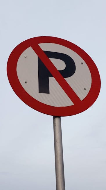 An empty parking lot situated in an urban area during daytime, with clear blue skies and scattered white clouds overhead. The asphalt surface of the parking area is marked with white lines designating individual parking spaces, some of which are occupied by small metal and glass bus shelters with green roofs positioned near the center and sides. Leafless trees line the perimeter of the lot, with a few bearing early spring blossoms, and tall lampposts with black fixtures are visible along the edges of the parking area. In the background, modern office or apartment buildings with large windows and balconies rise above the trees, indicating a city center setting. The parking lot is well-lit by natural sunlight, illustrating a typical scene where home relocation or furniture transport might involve careful planning for parking permissions, as managed by local authorities such as Hounslow Council, with services provided by companies like Man With a Van Hounslow for moves requiring parking permits and logistical support.