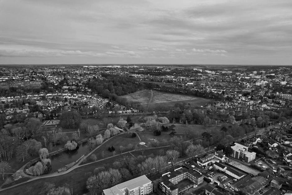 A black-and-white aerial photograph of a residential area near Hounslow Heath park, showing a spacious outdoor park with winding pathways, trees, and open grassy fields. Surrounding the park are rows of houses, apartment buildings, and commercial structures, with streets and parked cars visible along the roads. In the foreground, a property interior is visible with a furnished room that contains a wardrobe, cardboard packing boxes, a large plastic-wrapped item, and a moving trolley. The scene depicts the process of home relocation, including packing materials on the floor and the loading process into a nearby van, with a man from Man With a Van Hounslow carefully carrying or preparing furniture and boxes for transport, supporting furniture transport and packing during the move near Hounslow.