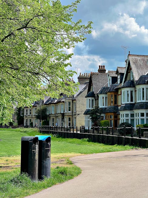 A wide outdoor scene at Hounslow Heath featuring a park with a gravel pathway leading towards a small body of water. To the left, there are tall, leafy green trees, including a prominent silver birch with white bark and drooping branches, and on the right, a large deciduous tree displaying vibrant orange and green foliage. In the foreground, a simple wooden park bench is positioned on the grass near the pathway. The area is bathed in soft afternoon light, casting gentle shadows. In the background, more trees and greenery extend along the water’s edge, creating a peaceful natural setting. This tranquil landscape exemplifies a typical backdrop for house removals or furniture transport services, with an open space suitable for loading and unloading items in proximity to the park and Hounslow’s natural environment, as provided by Man With a Van Hounslow.