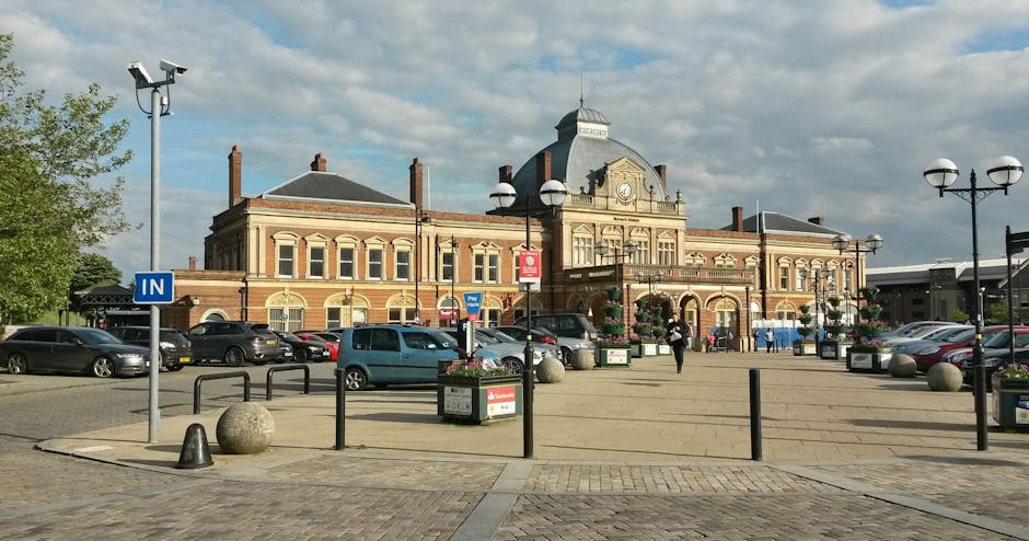 An exterior view of a historic brick building situated on Hounslow High Street, with a large paved open area in front used for parking and pedestrian access. Several cars are parked along the perimeter, including compact and family-sized vehicles, with some parked near lamp posts and bollards that outline the pedestrian zone. There are decorative street lamps with multiple globes, flower planters, and a blue 'IN' parking sign visible on the left. The building features arched windows, a central clock, intricate architectural details, and a dome roof, surrounded by modern amenities. The scene is illuminated by daytime natural light, with a partly cloudy sky overhead. This setting depicts a typical town centre environment suitable for home relocation or furniture transport, with visible parking and loading areas that could be utilized by a professional removals service like Man With a Van Hounslow during a house move or furniture transport process.