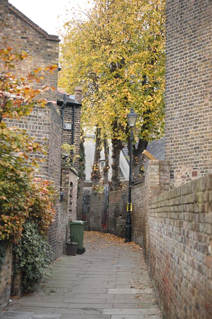 A narrow residential alleyway with a paved walkway surrounded by tall brick walls and buildings on both sides. On the left side, there are green shrubs and autumnal trees with yellow and orange leaves, some leaves fallen on the ground. A small green wheeled rubbish bin is positioned near the corner of the wall. On the right side, a tall brick wall runs parallel to the walkway. Midway down the alley, a vintage-style street lamp is mounted on a black pole with a yellow and black caution tape wrapped around its base. At the far end, a large deciduous tree with dense yellow foliage partially obscures a set of white residential buildings visible further back. The scene is illuminated by natural daylight, suggesting an autumn setting. This urban scene may relate to house removals and moving logistics, possibly illustrating a tight access point evaluated during home relocation or furniture transport, as part of professional services offered by Man With a Van Hounslow.