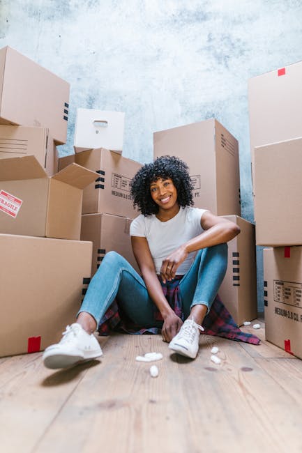 A young woman with curly dark hair, wearing a white T-shirt, blue jeans, and white sneakers, is sitting on a wooden floor surrounded by multiple cardboard boxes of various sizes, some sealed with red tape and others partially open, in an indoor space with a textured blue wall. She is smiling at the camera and appears to be in the process of packing or organizing belongings for a house removal. Several foam packing peanuts are scattered on the floor in front of her. The boxes are stacked and placed around her, indicating an active packing or furniture transport process, typical of home relocation services provided by companies like Man With a Van Hounslow. The setting captures the routine scene of packing during a move, with the focus on the physical effort involved in packing and preparing items for transport.