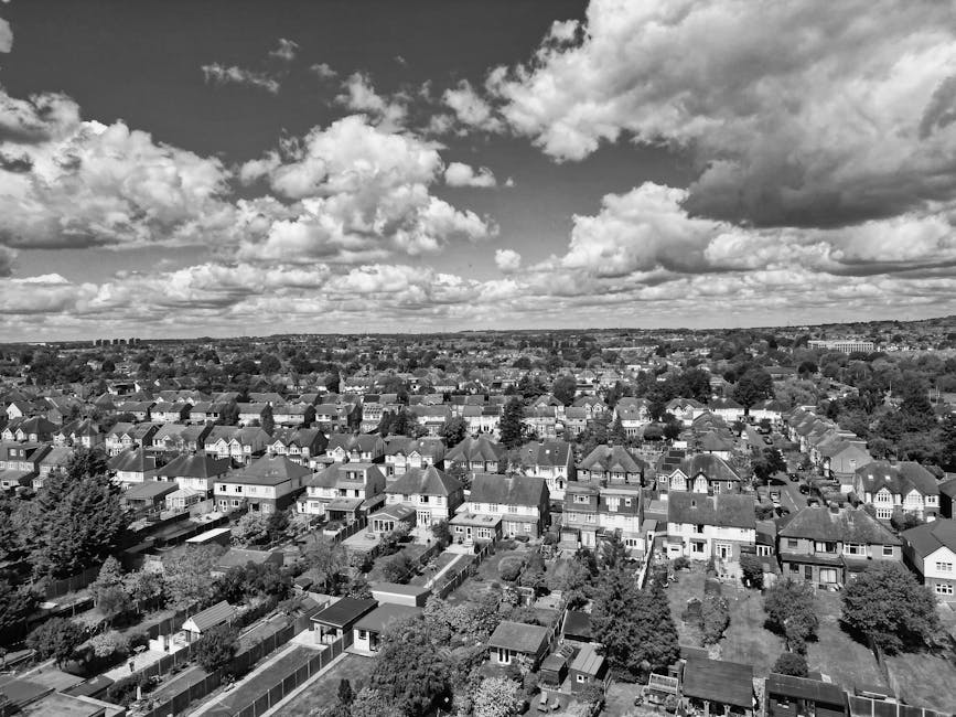 Black and white aerial photograph of a suburban residential area during daytime, showing numerous houses with pitched roofs arranged in a neat grid pattern. Some houses have small gardens enclosed by