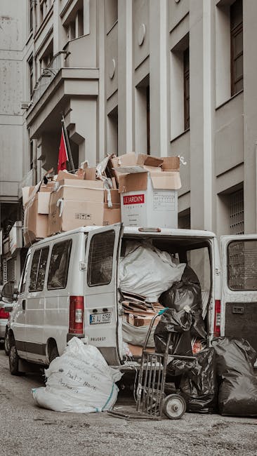 A white commercial van parked on a city street in front of a multi-storey building, with its rear doors open revealing a loading area filled with various packing materials and household items. Inside the van, there are large black rubbish bags, cardboard boxes, and wrapped furniture parts, indicating a home relocation or removal process. Some boxes are stacked on top of each other towards the vehicle’s roof, secured with straps or placed directly inside the van. Outside, a small hand truck or dolly is positioned next to the open rear doors, suggesting the loading or unloading of furniture and boxes. The building's facade is light-colored with several windows and architectural details, contributing to an urban environment typical for house removals performed by companies like Man With a Van Hounslow. The scene captures the logistical phase of packing and transporting belongings, highlighting professional furniture transport and moving services.
