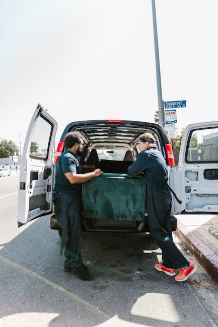 Two movers dressed in dark clothing and safety glasses are loading a large, dark green wrapped piece of furniture into the rear of a white van parked on a paved street. The van's rear doors are wide open, revealing the spacious interior designed for furniture transport. The scene takes place outdoors in daylight, with a clear sky visible above, and features nearby street signage including a blue sign attached to a pole. The pavement and curb are visible in the foreground, and in the background, there is a supermarket or commercial building with parking areas. The furniture appears to be carefully wrapped in protective fabric or padding as part of packing and moving procedures, supporting a home relocation or furniture transport service offered by Man With a Van Hounslow, as indicated by the context of house removals and relocation logistics.