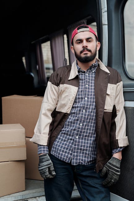 A man with a beard and wearing a red cap, plaid shirt, and a beige and brown work jacket stands inside the back of a van used for house removals, with cardboard boxes stacked behind him on the van's floor. The van's interior features dark trim and a window on the side, and the man is wearing gloves, suggesting he is involved in furniture transport or packing during a home relocation. The environment is lit naturally, highlighting the loading process associated with moving services provided by Man With a Van Hounslow, as seen on the page about urgent same-day removal services in Hounslow.