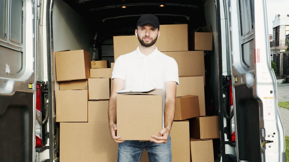 A man wearing a white shirt, dark cap, and jeans is standing at the open rear of a moving van, holding a cardboard box. The van's interior is filled with neatly stacked cardboard boxes of various sizes, some wrapped in plastic and others unwrapped, arranged to facilitate efficient loading and transport during a home relocation. The loading process appears organized, with additional boxes on the pavement just outside the van, indicating active packing and moving activities. A section of the street and residential buildings with outdoor balconies can be seen in the background, outside the property where the loading is taking place. The scene is well-lit, showing a typical moving day operation, with visible equipment such as moving blankets and straps possibly used for securing furniture and boxes during transit. This image exemplifies the services provided by Man With a Van Hounslow, specializing in removals and furniture transport within the Hounslow area.