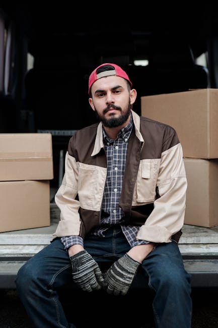 A man with a beard and wearing a red cap, plaid shirt, and a beige and brown work jacket stands inside the back of a van used for house removals, with cardboard boxes stacked behind him on the van's floor. The van's interior features dark trim and a window on the side, and the man is wearing gloves, suggesting he is involved in furniture transport or packing during a home relocation. The environment is lit naturally, highlighting the loading process associated with moving services provided by Man With a Van Hounslow, as seen on the page about urgent same-day removal services in Hounslow.